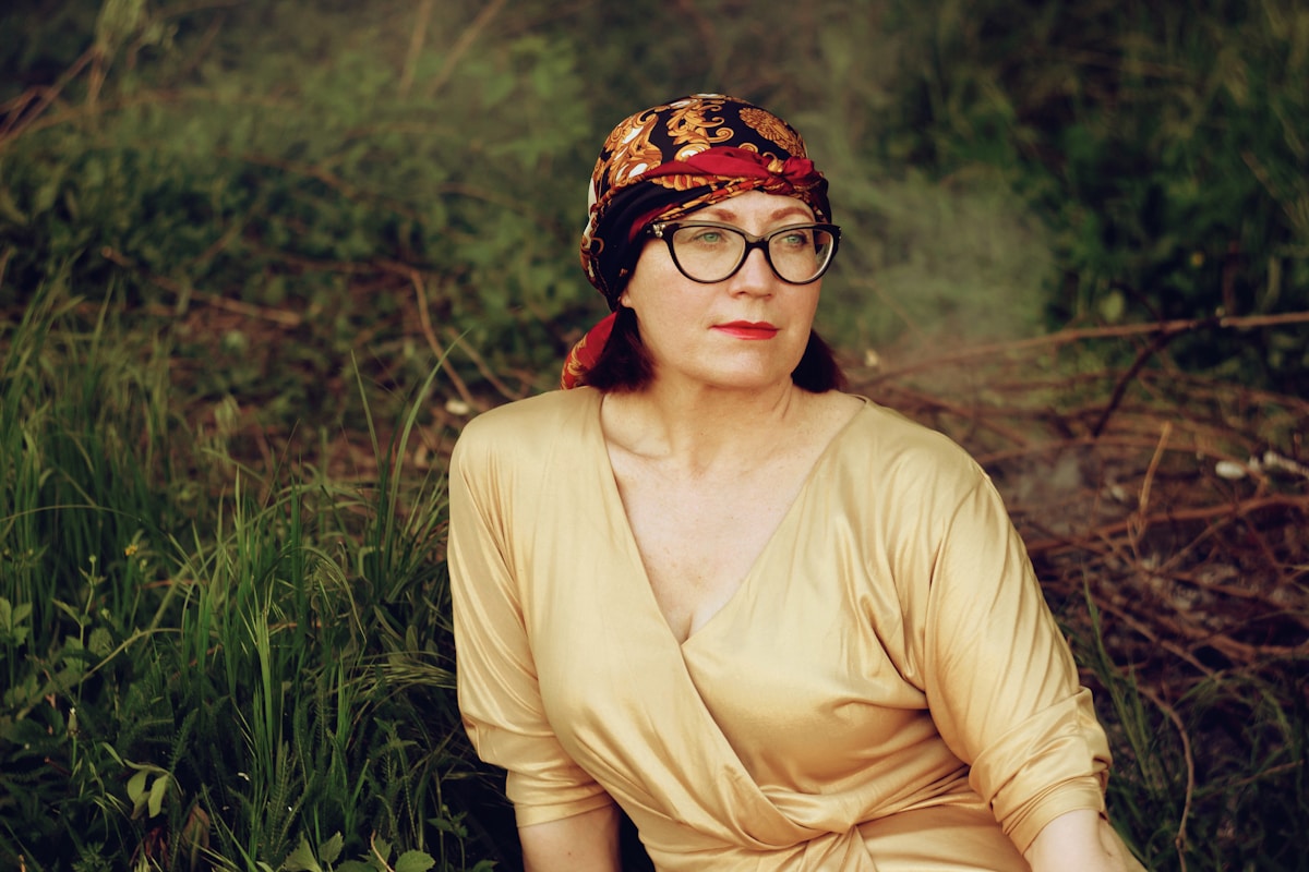 Femme âgée, portant un foulard coloré et des lunettes, assise dans un environnement naturel.